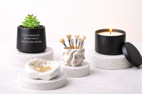 Self-care gift set on marble pedestals featuring a black succulent jar with message, a face brush holder, a hand trinket dish, and a black candle.