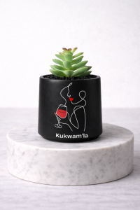 Matte black mini succulent jar with a green succulent and minimalist line art of a woman holding a red wine glass, with the text “Kukwam’la”, displayed on a white marble pedestal.