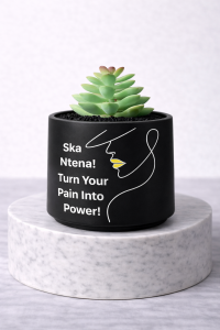 Matte black mini succulent jar with line art face design and the message “Ska Ntena! Turn Your Pain Into Power!” displayed on a white marble pedestal against a clean light grey background.